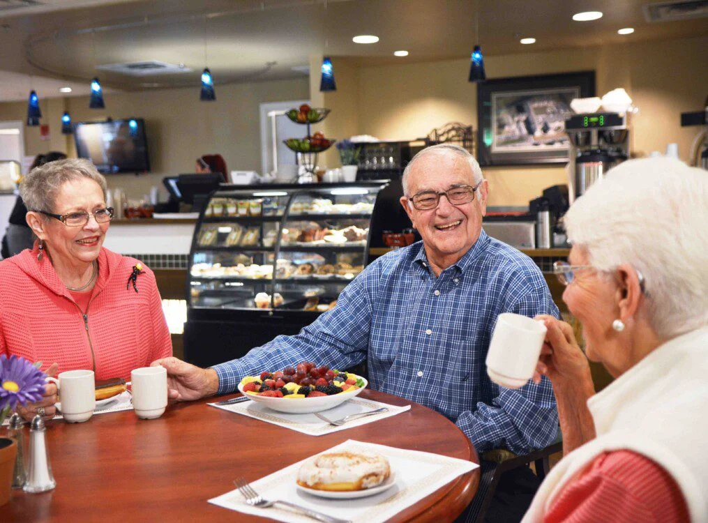 smiling group of senior friends enjoy a meal at a cafe-style venue in their senior living community