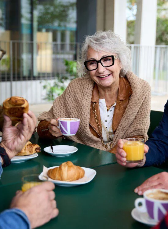 Mature grey haired woman smiling enjoying coffee with group friends on terrace of old people home.