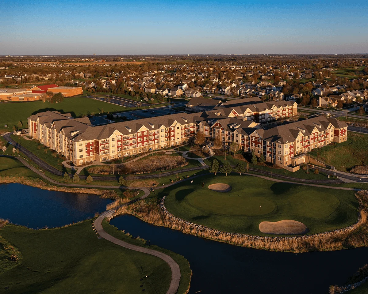 Aerial view of residential buildings surrounded by greenery and water features near suburban homes under clear sky.