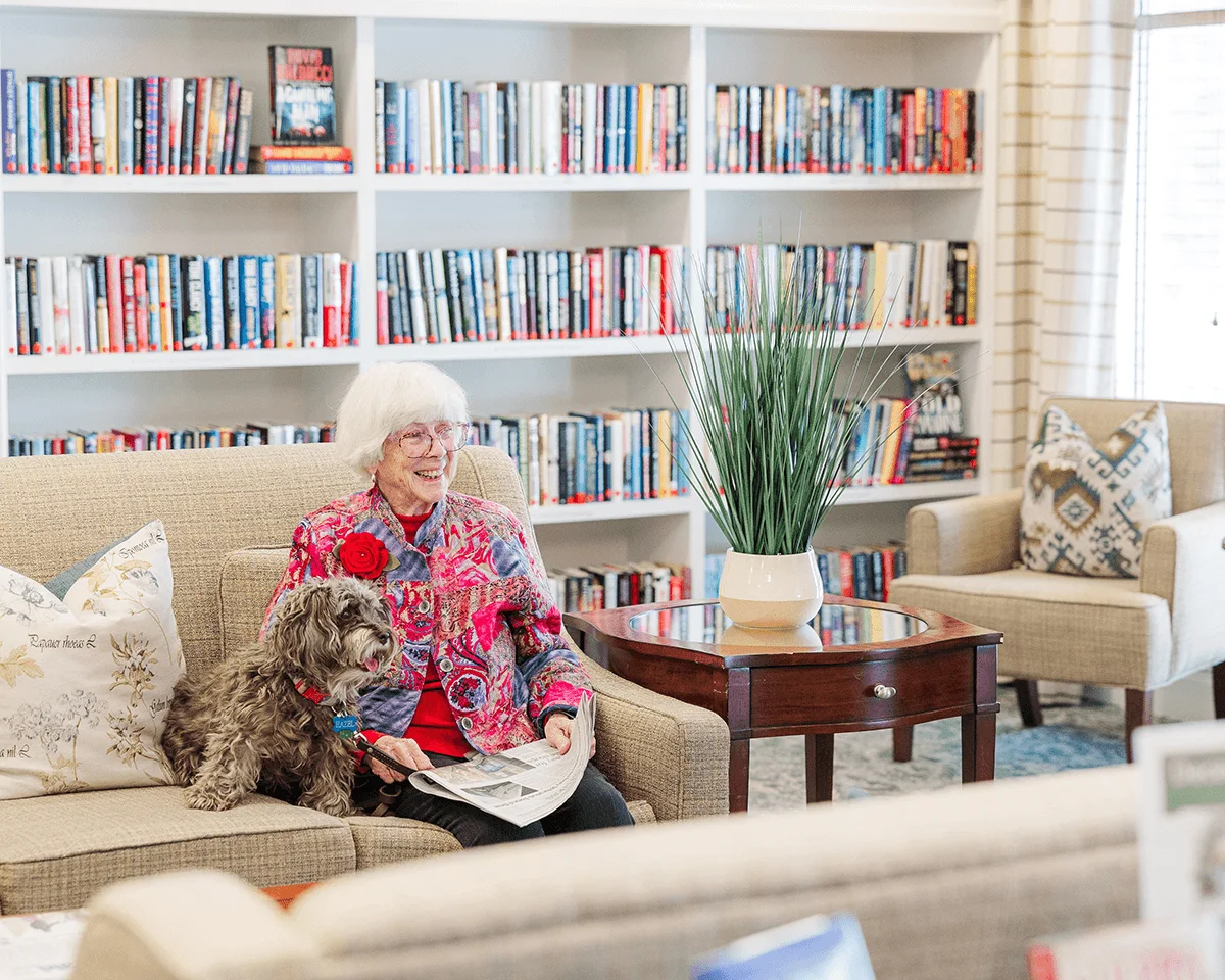 Woman reading with dog on cozy couch near bookshelves and coffee table.