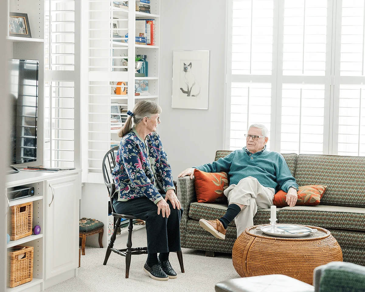A cozy living room with two people conversing near a couch and bookshelves.