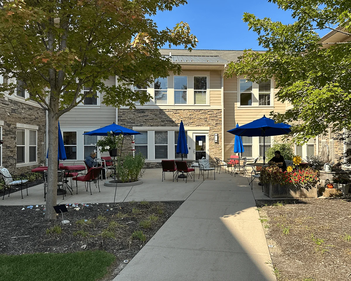 A sunny courtyard with blue umbrellas and red chairs under trees near stone buildings on a clear day.