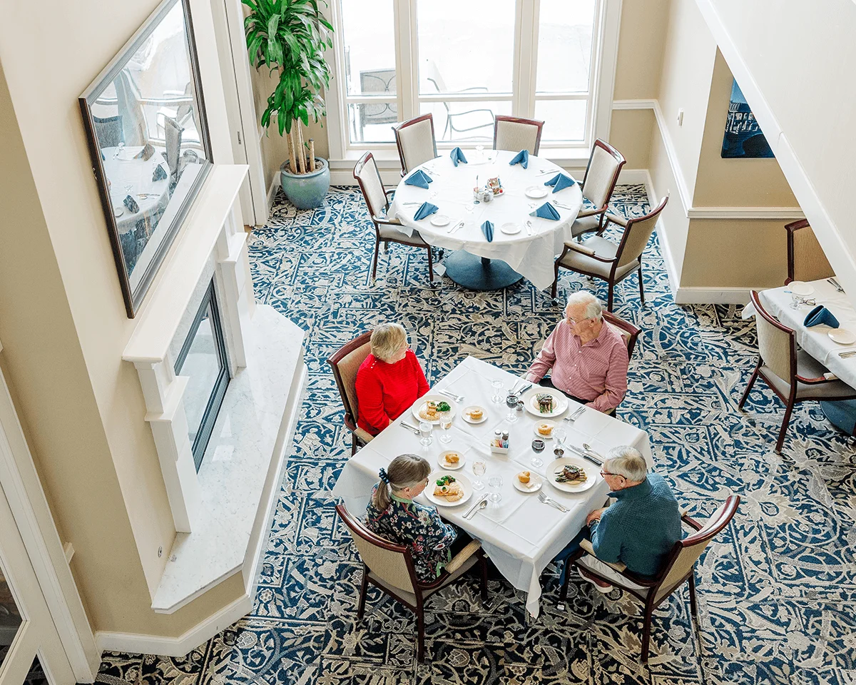 Four people enjoy a meal at tables with white tablecloths and blue napkins in an elegant dining room setting.