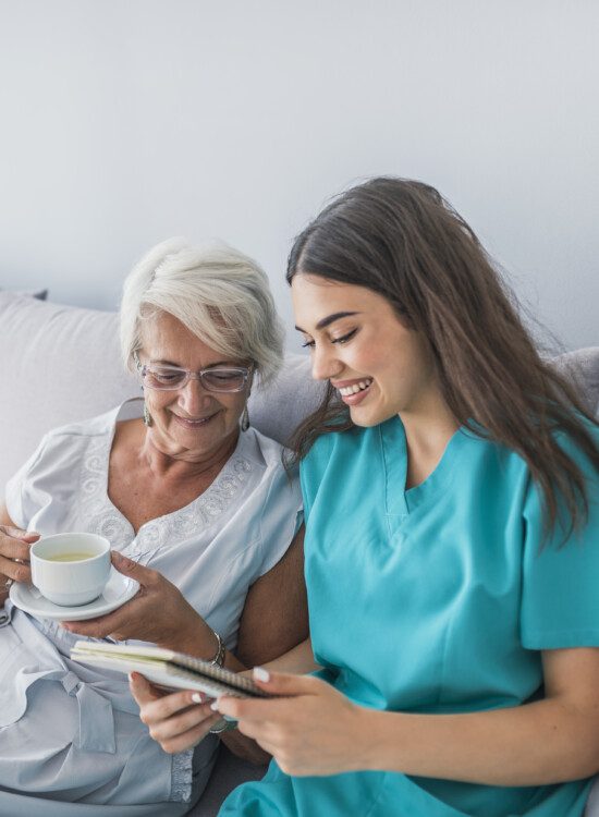 Happy patient is holding caregiver for a hand while spending time together. Elderly woman in nursing home and nurse. Aged elegant woman and tea time at nursing home