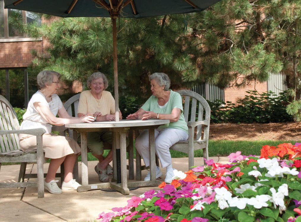 group of seniors enjoy a meal together at a bistro-style table outdoors at their senior living community, surrounded by flowers