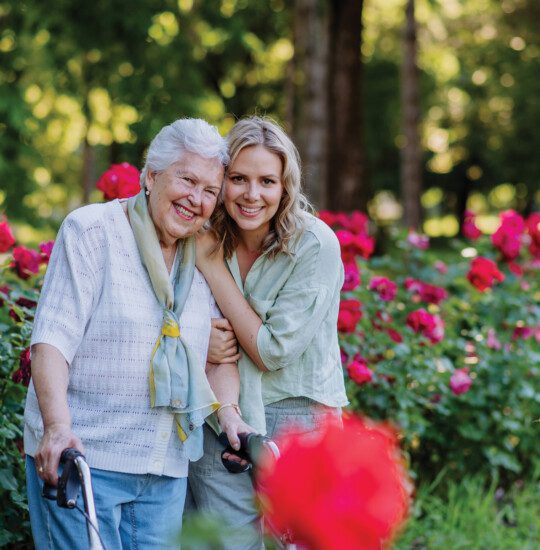 Portrait of adult granddaughter with senior grandmother on walk in park, with roses at background